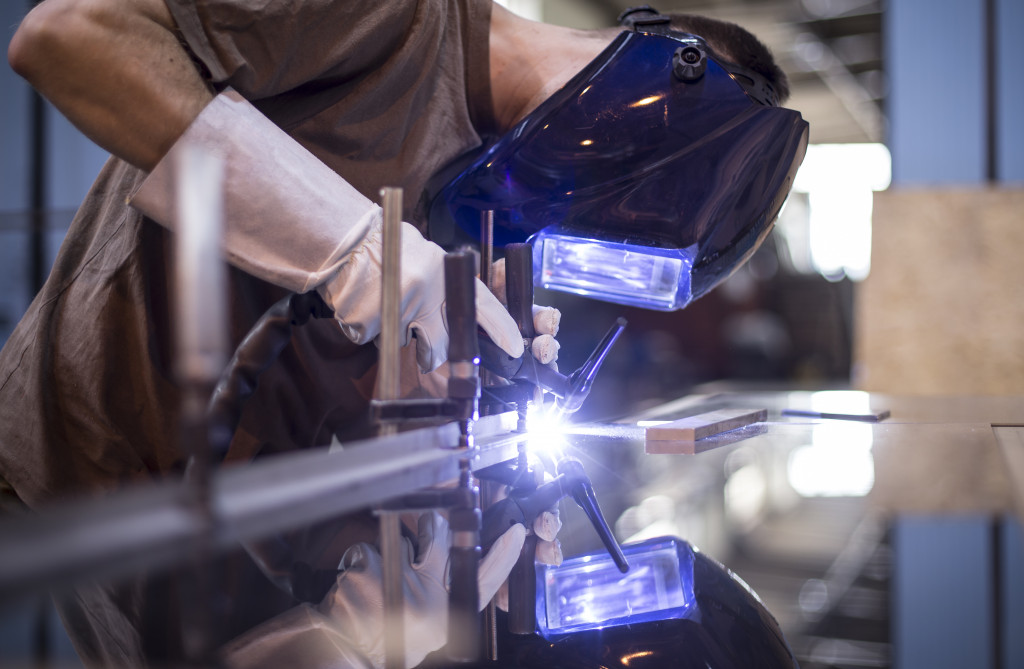 A worker wearing a welding mask and gloves uses a TIG welder on a large sheet of metal
