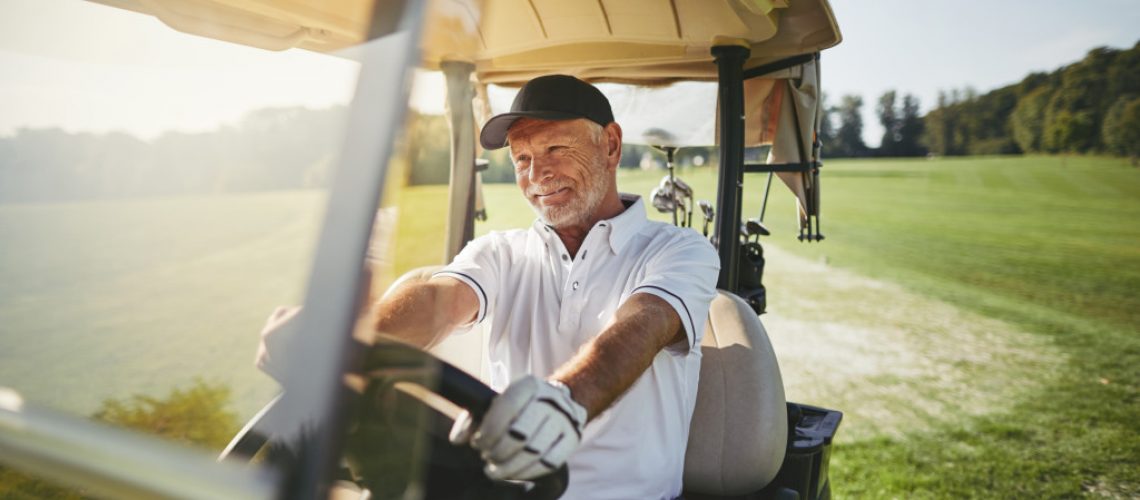 An older man wearing gloves and golfer attire smiling while driving a golf cart in a golf club fairway