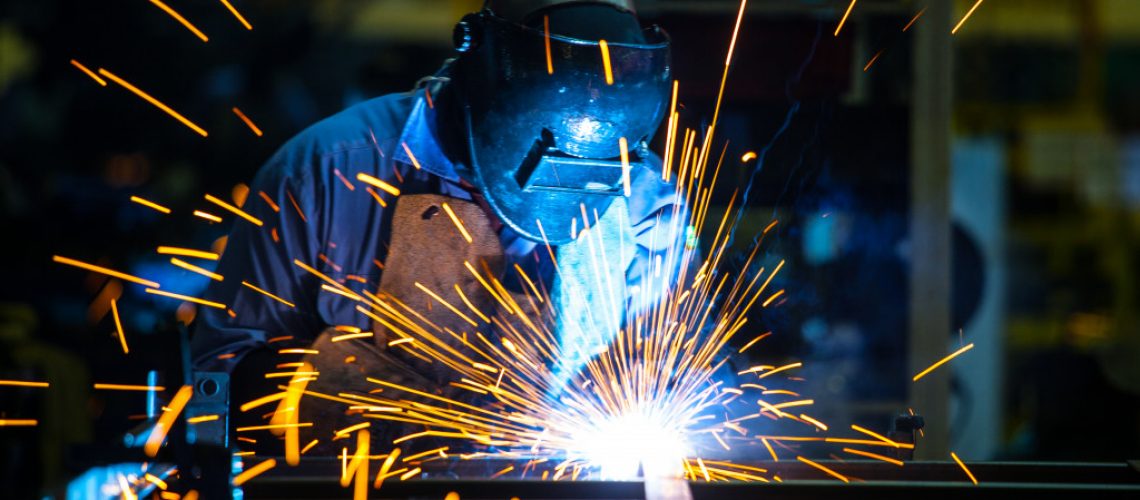 Bright sparks erupt in front of a welder wearing a welding mask, apron, and gloves